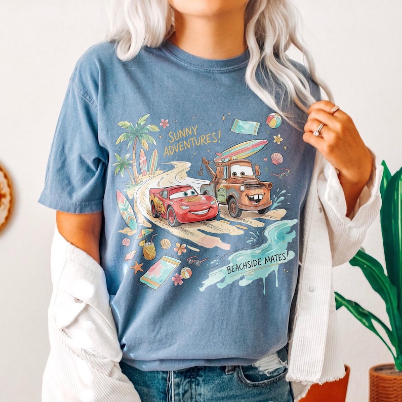 May include: A blue short-sleeve t-shirt featuring a graphic print of Lightning McQueen and Mater driving on a beach road. The design includes palm trees, surfboards, flowers, and the text Sunny Adventures and Beachside Mates in yellow and black.
