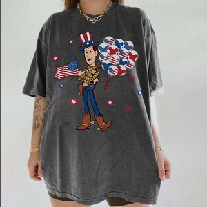 May include: Dark grey t-shirt with a cartoon cowboy character holding a US flag and balloons with patriotic designs. Red, white, and blue stars are scattered around the graphic. The shirt has short sleeves and a relaxed fit.