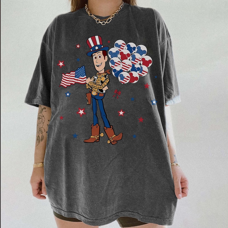 May include: Dark grey t-shirt with a cartoon cowboy character holding a US flag and balloons with patriotic designs. Red, white, and blue stars are scattered around the graphic. The shirt has short sleeves and a relaxed fit.