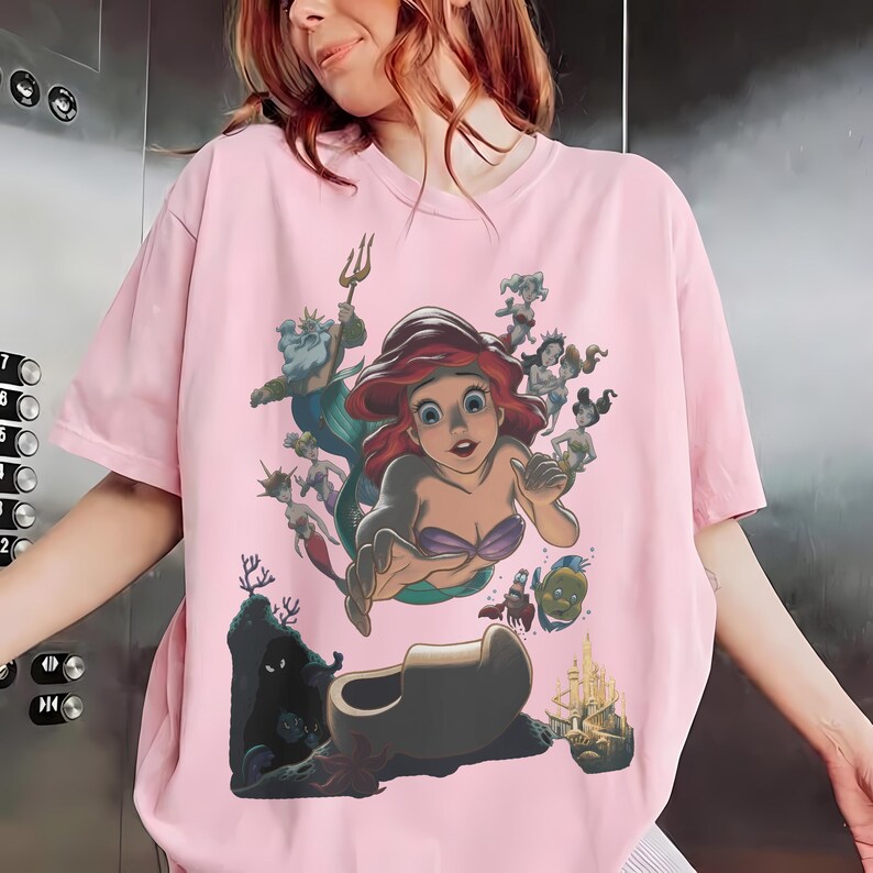 May include: A light pink t-shirt featuring a graphic print of Ariel from The Little Mermaid, surrounded by King Triton, Flounder, Sebastian, and other underwater characters, with a castle and coral reef depicted at the bottom of the design.