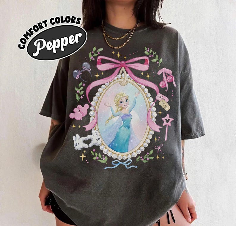 May include: A charcoal gray t-shirt featuring a graphic print of a blonde character in a blue dress inside a pearl-bordered oval frame. The design includes a pink bow, white cartoon gloves forming a heart, and small star and cherry accents.