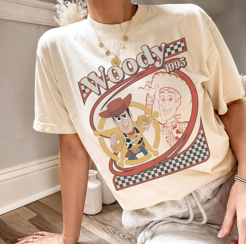 May include: Cream-colored t-shirt featuring a graphic of Woody from Toy Story. The design includes Woody in a cowboy hat, the word "Woody" in red, and the year "1995". The shirt has short sleeves and a relaxed fit.