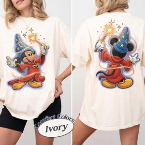 May include: Ivory t-shirt with front and back graphics of Mickey Mouse as a sorcerer. Mickey is in a blue wizard hat and red robe, holding a wand. The shirt is a Comfort Colors brand.