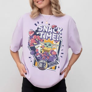 May include: A light purple short-sleeve t-shirt featuring a graphic print of a green creature eating snacks. The text reads SNACK TIME! and Bounty Hunters. The design includes purple and pink cloud-like shapes and a helmet icon in a frame.