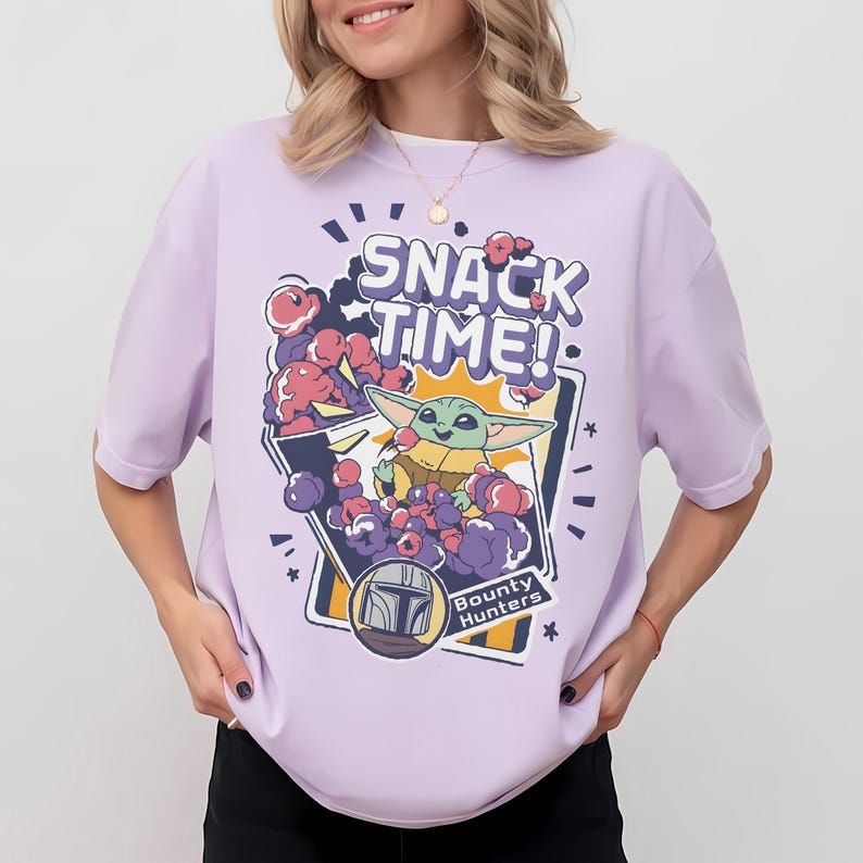 May include: A light purple short-sleeve t-shirt featuring a graphic print of a green creature eating snacks. The text reads SNACK TIME! and Bounty Hunters. The design includes purple and pink cloud-like shapes and a helmet icon in a frame.