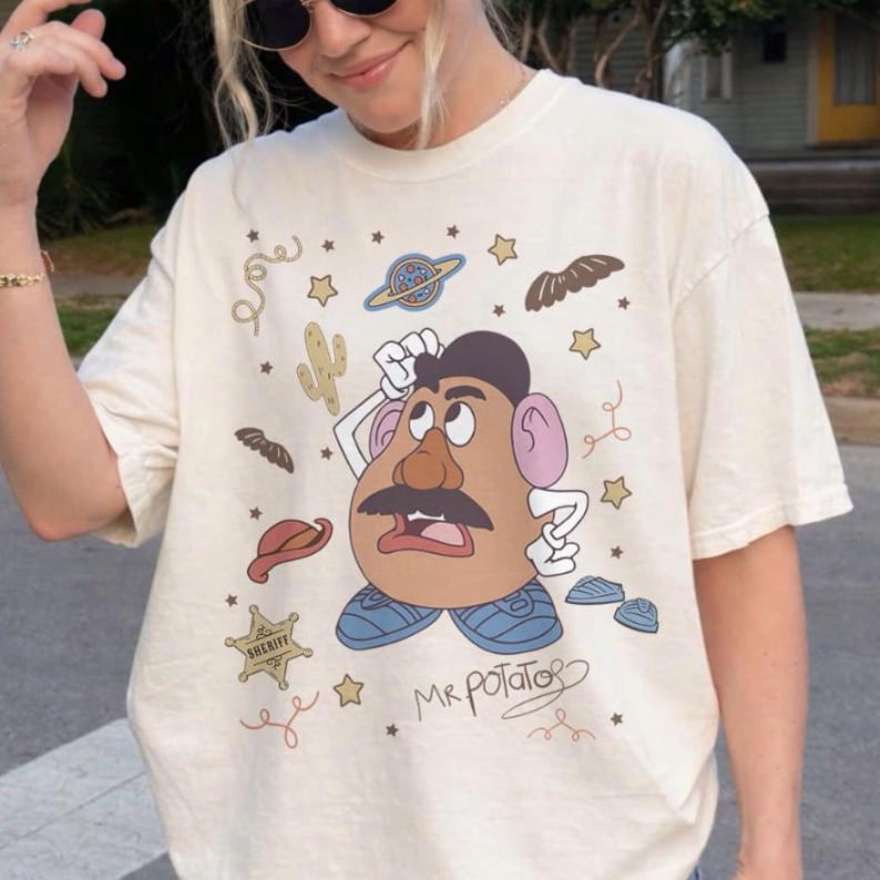 May include: Cream-colored t-shirt featuring a cartoon Mr. Potato Head character with a large mustache, wearing blue shoes. The design includes a sheriff's badge, stars, and a cactus. The words "Mr. Potato" are written in a whimsical font.