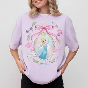 May include: A light purple t-shirt featuring a central oval frame decorated with pearls and a pink bow. Inside the frame is an illustration of a blonde character in a blue dress. Surrounding the frame are small icons including mouse ears, cherries, balloons, and a star wand.