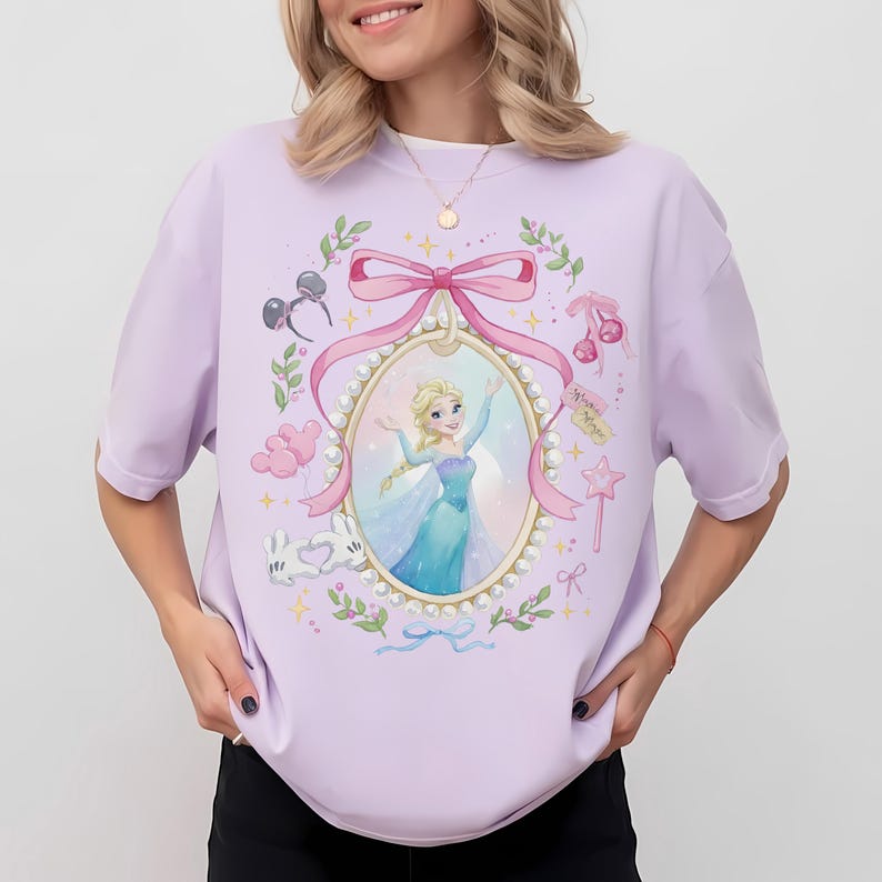 May include: A light purple t-shirt featuring a central oval frame decorated with pearls and a pink bow. Inside the frame is an illustration of a blonde character in a blue dress. Surrounding the frame are small icons including mouse ears, cherries, balloons, and a star wand.