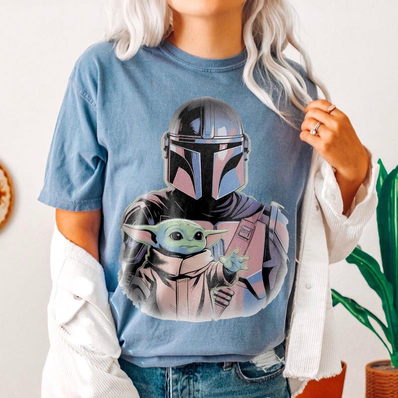 May include: A light blue t-shirt featuring a graphic of The Mandalorian and Grogu. The Mandalorian is depicted in his helmet and armor, with Grogu reaching out. The graphic has a comic book style with pink and purple accents.