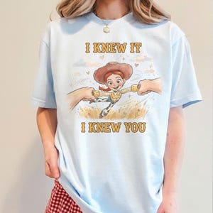 May include: A light blue t-shirt featuring a graphic of a cartoon cowgirl character with red hair and a brown hat, being held by two hands. The text "I KNEW IT" appears above the character and "I KNEW YOU" appears below in yellow lettering.