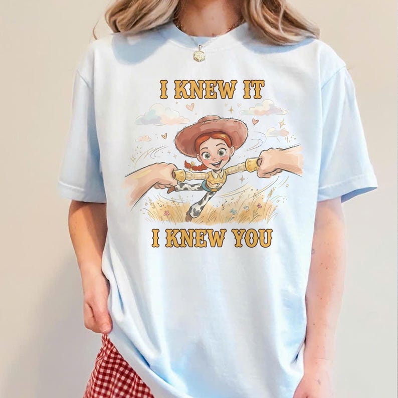 May include: A light blue t-shirt featuring a graphic of a cartoon cowgirl character with red hair and a brown hat, being held by two hands. The text "I KNEW IT" appears above the character and "I KNEW YOU" appears below in yellow lettering.