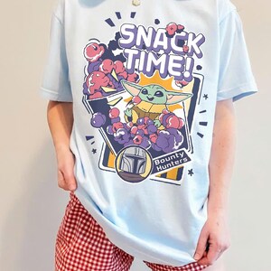 May include: A light blue t-shirt featuring a graphic print of a small green creature eating purple snacks from a bag. The text SNACK TIME! appears at the top, and a circular emblem with a helmet icon and the words Bounty Hunters is below.