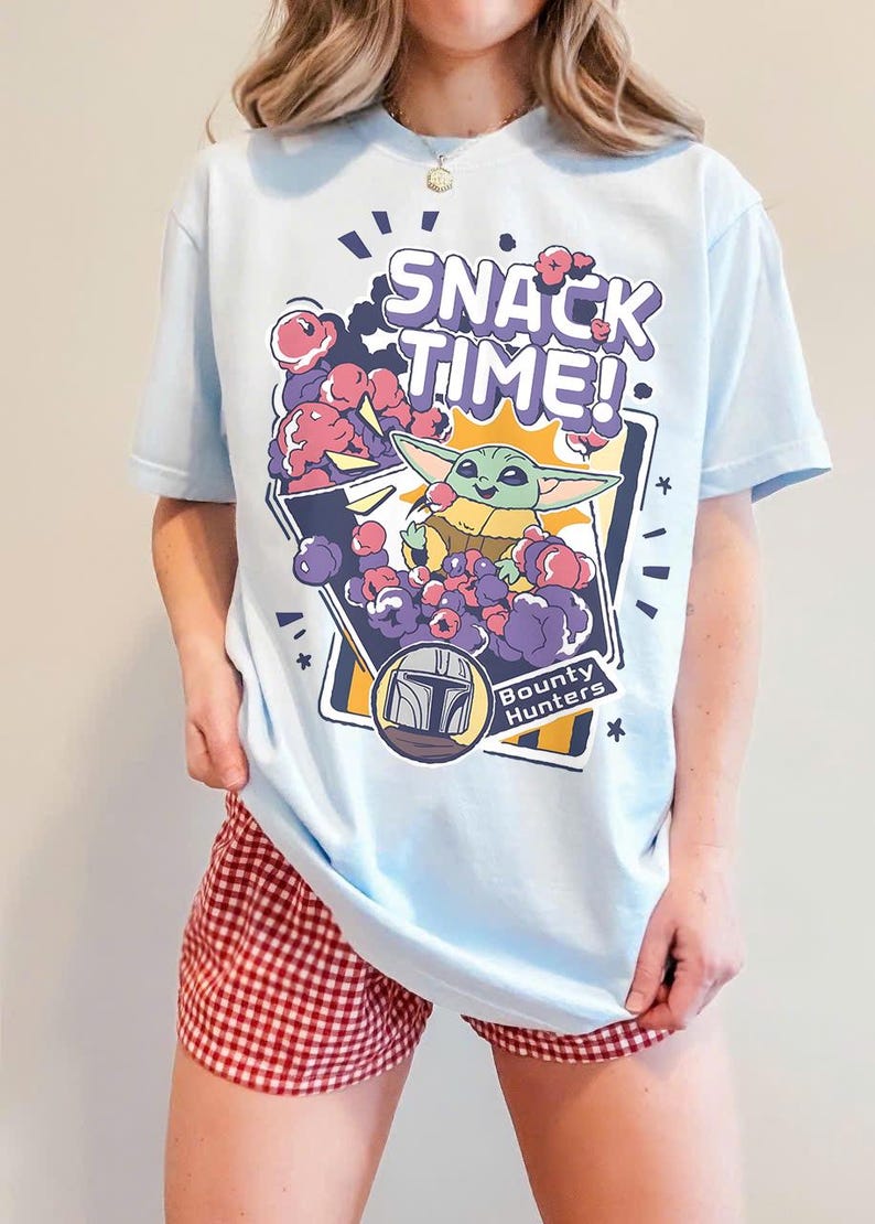 May include: A light blue t-shirt featuring a graphic print of a small green creature eating purple snacks from a bag. The text SNACK TIME! appears at the top, and a circular emblem with a helmet icon and the words Bounty Hunters is below.