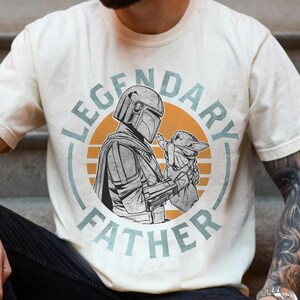 K&ouml;nnte beinhalten: Cremefarbenes T-Shirt mit einem kreisf&ouml;rmigen Aufdruck. Der Aufdruck zeigt eine Figur in R&uuml;stung, die eine kleine Kreatur h&auml;lt, mit dem Text "LEGENDARY FATHER". Das Design verwendet Grau-, Orange- und Blaut&ouml;ne.