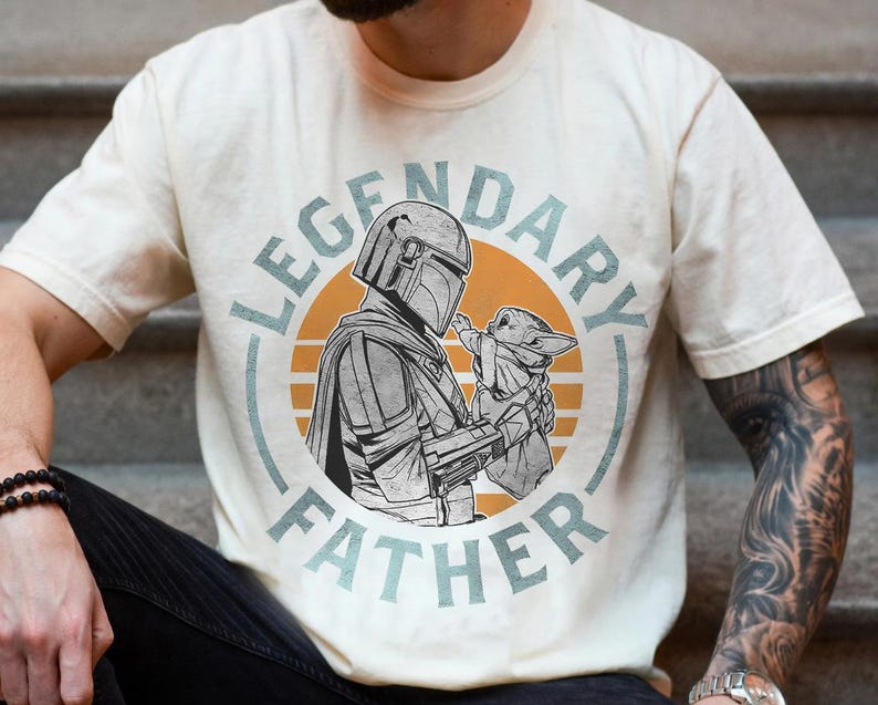 K&ouml;nnte beinhalten: Cremefarbenes T-Shirt mit einem kreisf&ouml;rmigen Aufdruck. Der Aufdruck zeigt eine Figur in R&uuml;stung, die eine kleine Kreatur h&auml;lt, mit dem Text "LEGENDARY FATHER". Das Design verwendet Grau-, Orange- und Blaut&ouml;ne.