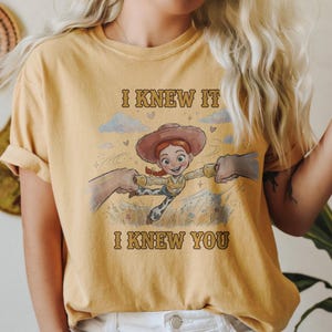May include: A mustard yellow t-shirt featuring a graphic print of a cowgirl character with red hair and a brown hat. The text "I KNEW IT" appears above the character and "I KNEW YOU" appears below, with illustrations of clouds and grass.