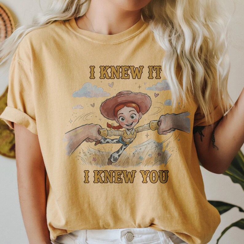 May include: A mustard yellow t-shirt featuring a graphic print of a cowgirl character with red hair and a brown hat. The text "I KNEW IT" appears above the character and "I KNEW YOU" appears below, with illustrations of clouds and grass.