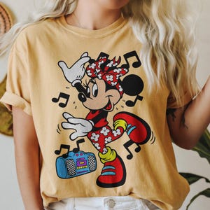 Pu&ograve; includere: T-shirt giallo senape con una grafica di Minnie Mouse che balla. Minnie indossa un completo rosso con dettagli floreali bianchi, un fiocco abbinato e scarpe rosse. Un boombox blu e note musicali completano il design.