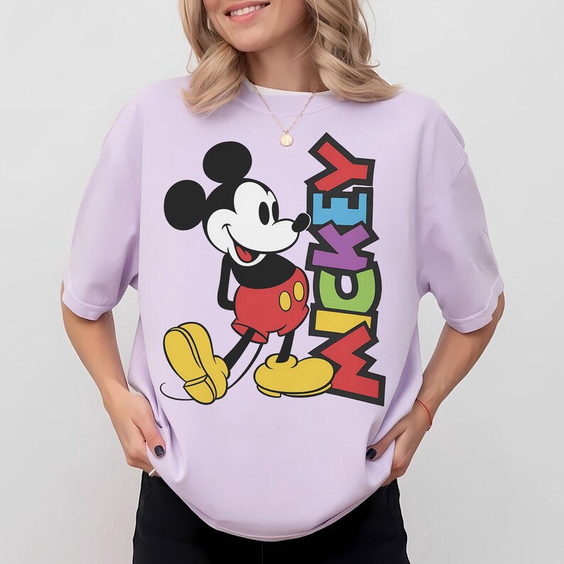 May include: A light purple t-shirt featuring a graphic of Mickey Mouse in his classic pose. Mickey is wearing red shorts, yellow shoes, and a black top. The word "MICKEY" is printed vertically in colorful letters.