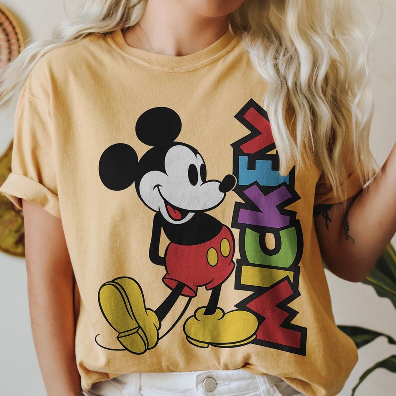 May include: Mustard yellow t-shirt featuring a smiling Mickey Mouse graphic. Mickey is depicted in his classic attire: red shorts, yellow shoes, and white gloves. The word "Mickey" is printed in colorful, stacked letters next to the character.