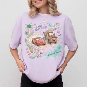 May include: A light purple t-shirt featuring a colorful graphic of Lightning McQueen and Mater from the movie Cars driving on a beach road. The design includes palm trees, surfboards, beach towels, and the text Sunny Adventures and Beachside Mates.