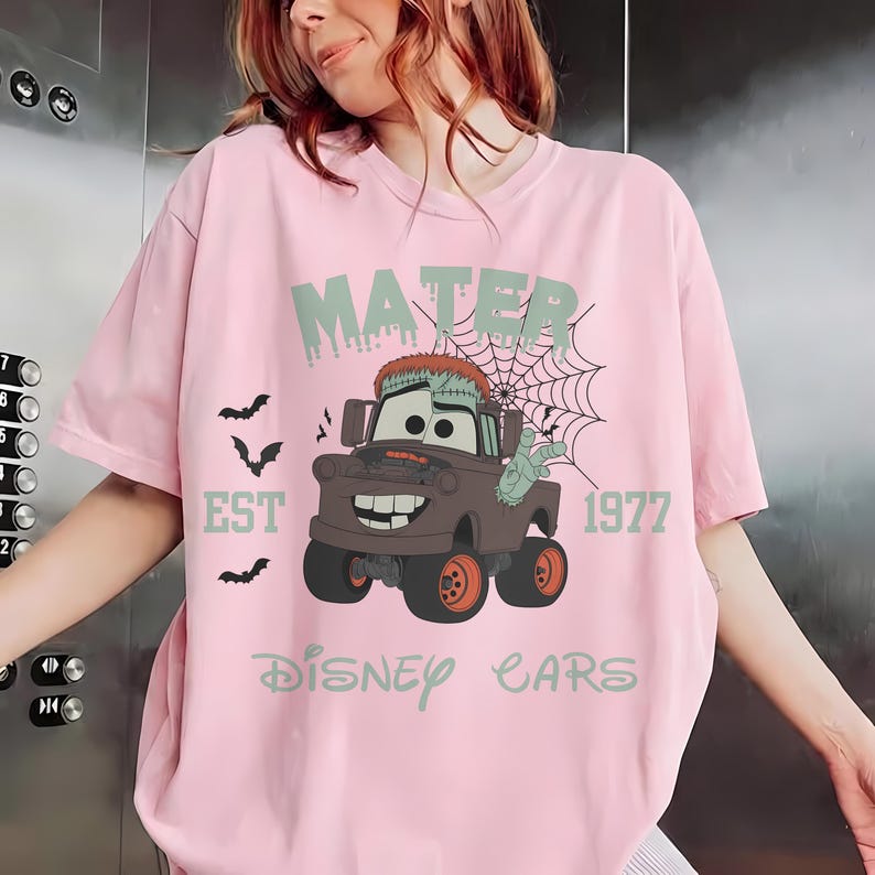 May include: Pink t-shirt featuring a cartoon Mater from Disney Cars, with a Halloween theme. The design includes the text "MATER EST 1977" and a spiderweb. The truck is brown with orange wheels and a zombie-like appearance. The words "Disney Cars" are also printed on the shirt.