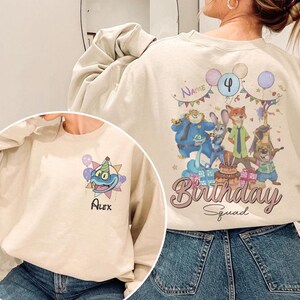 K&ouml;nnte beinhalten: Beiges Sweatshirt mit farbenfroher Cartoon-Grafik mit Figuren, Luftballons und dem Text "Birthday Squad". Die Zahl "4" wird angezeigt. Der Name "Alex" ist ebenfalls sichtbar.