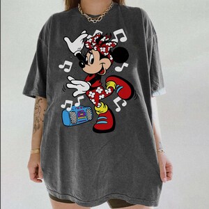 Pu&ograve; includere: T-shirt grigio scuro con grafica di Minnie Mouse che balla. Minnie indossa un completo rosso con dettagli floreali bianchi, scarpe rosse e un fiocco rosso. &Egrave; presente anche un boombox blu e note musicali.