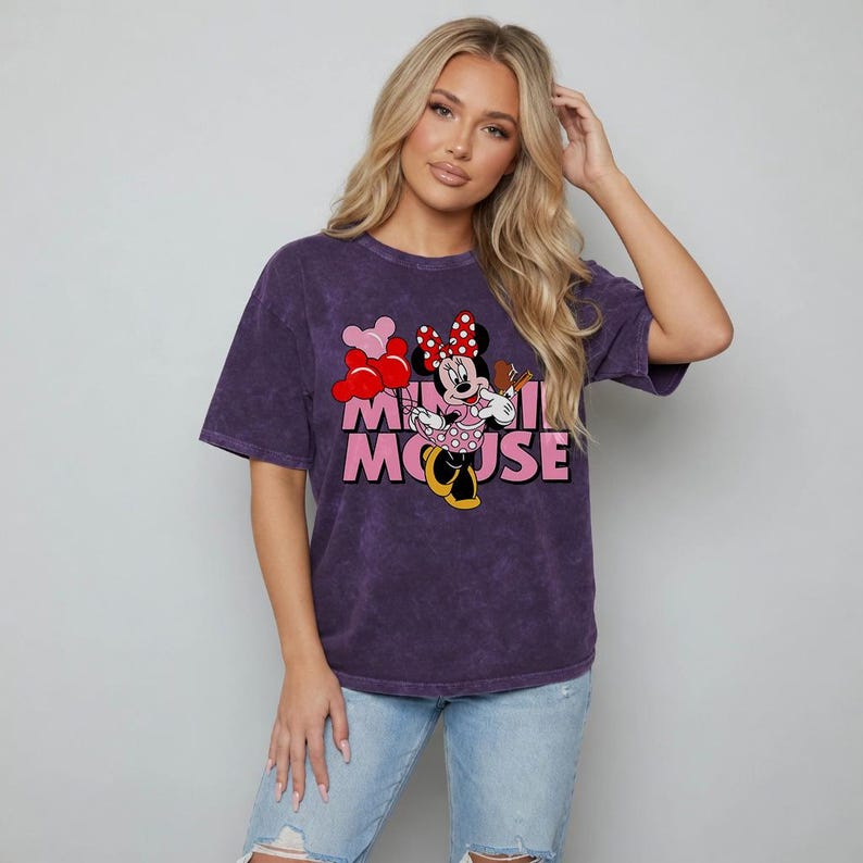 May include: A purple t-shirt featuring a graphic of Minnie Mouse holding balloons and a musical instrument. The text "MINNIE MOUSE" is printed below the character in pink. The shirt has short sleeves and a crew neck.