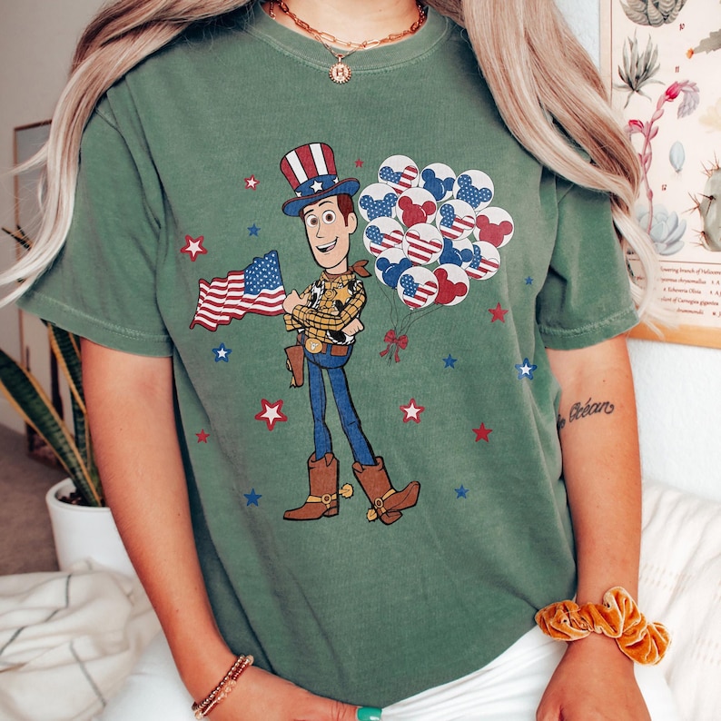 May include: Green t-shirt with a cartoon illustration of Woody from Toy Story, holding a US flag and balloons. The balloons have red, white, and blue patterns. Red and blue stars are scattered around the design.