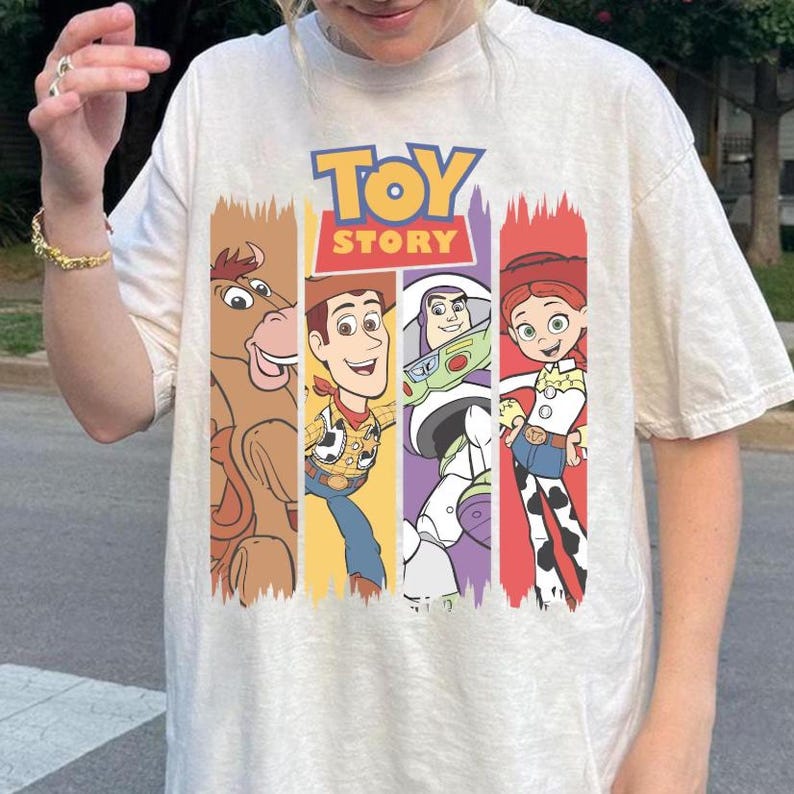 May include: Cream-colored t-shirt featuring the "Toy Story" logo and character illustrations. The design includes images of Woody, Buzz Lightyear, Jessie, and Bullseye, set against colorful vertical stripes.