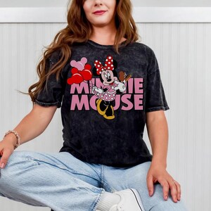May include: Black t-shirt featuring Minnie Mouse holding balloons and a treat. The graphic includes the text "MINNIE MOUSE" in pink. The shirt has a vintage wash appearance and is paired with light blue jeans.