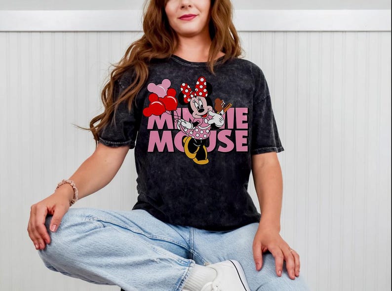 May include: Black t-shirt featuring Minnie Mouse holding balloons and a treat. The graphic includes the text "MINNIE MOUSE" in pink. The shirt has a vintage wash appearance and is paired with light blue jeans.