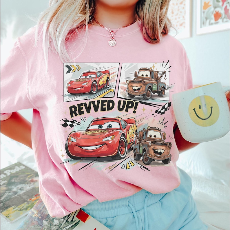 May include: A light pink t-shirt featuring a graphic print of the characters Lightning McQueen and Mater from the movie Cars. The design includes the text REVVED UP! along with checkered flag motifs and comic-style panels showing the characters.