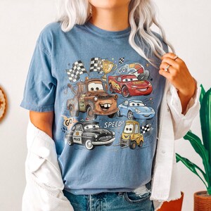 May include: A blue short-sleeve t-shirt featuring a graphic print of various animated car characters, including a brown tow truck, a red race car, a blue sports car, a black police car, and a yellow forklift, with the text GO! SPEED!.