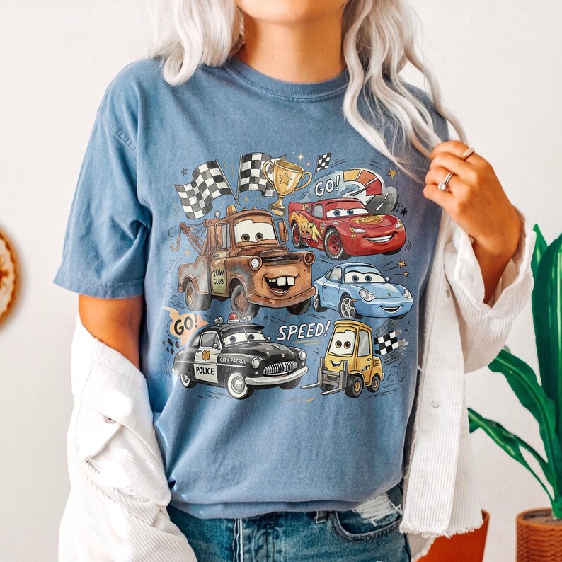 May include: A blue short-sleeve t-shirt featuring a graphic print of various animated car characters, including a brown tow truck, a red race car, a blue sports car, a black police car, and a yellow forklift, with the text GO! SPEED!.