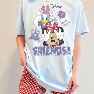 Może przedstawiać: Jasnoniebieska koszulka z Daisy Duck i Minnie Mouse z Disneya ze słowem "FRIENDS!" w kolorze fioletowym. Koszulka ma dodatkowy tekst z napisem "You Are Cool" i "You Are Amazing".
