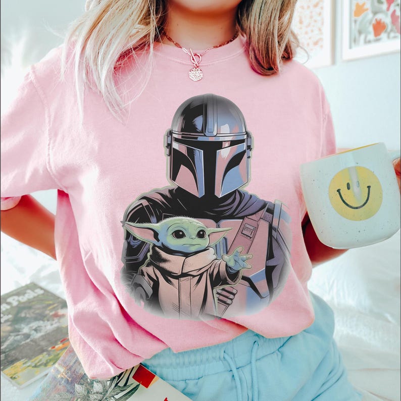 May include: Pink t-shirt featuring a graphic of The Mandalorian and Grogu. The Mandalorian is in his helmet and armor, with Grogu reaching out. A white mug with a yellow smiley face is held in the hand.