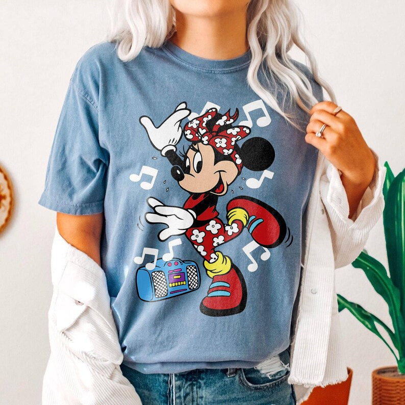Pu&ograve; includere: Maglietta blu con Minnie Mouse che balla, circondata da note musicali. Minnie Mouse indossa un completo floreale rosso e bianco e scarpe rosse. Un boombox blu &egrave; presente nel design.