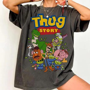 Pode incluir: T-shirt cinzenta escura com um gr&aacute;fico de desenho animado com o texto "Thug Story". O gr&aacute;fico inclui v&aacute;rios personagens de desenhos animados, como um dinossauro, Woody, Buzz Lightyear e outros, com acess&oacute;rios tem&aacute;ticos de dinheiro e g&acirc;ngsteres.