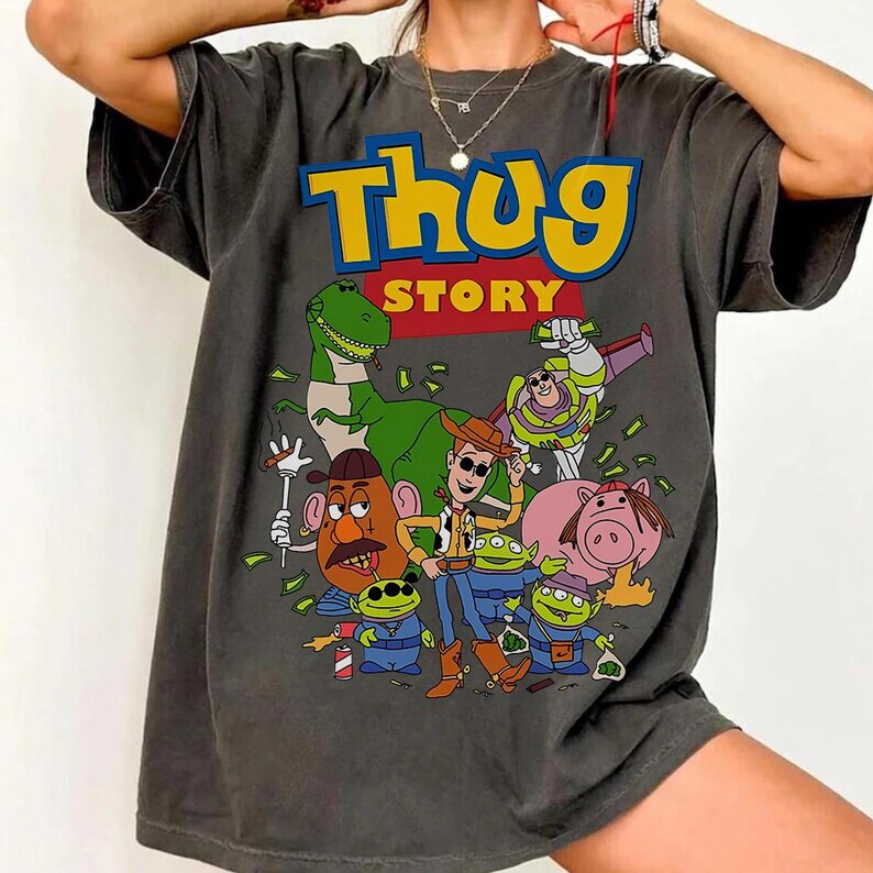 Pode incluir: T-shirt cinzenta escura com um gr&aacute;fico de desenho animado com o texto "Thug Story". O gr&aacute;fico inclui v&aacute;rios personagens de desenhos animados, como um dinossauro, Woody, Buzz Lightyear e outros, com acess&oacute;rios tem&aacute;ticos de dinheiro e g&acirc;ngsteres.