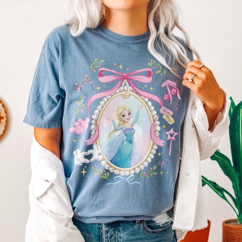 May include: A blue short-sleeve t-shirt featuring a graphic of a blonde character in a blue dress inside a pearl-bordered oval frame. The design includes a large pink bow, small pink mouse-ear silhouettes, a star wand, and green leaves.