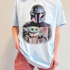 May include: Light blue t-shirt featuring a graphic of The Mandalorian and Grogu. The Mandalorian is wearing his helmet and armor, and Grogu is reaching out. The shirt is a light blue color.