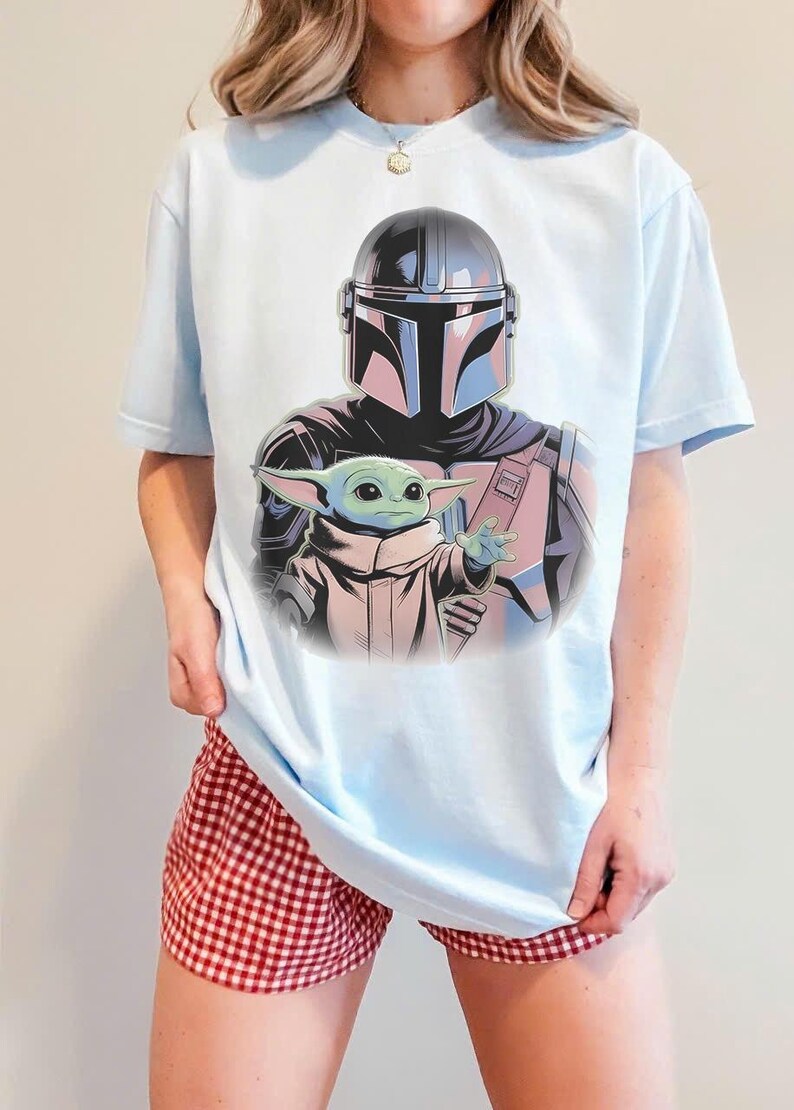 May include: Light blue t-shirt featuring a graphic of The Mandalorian and Grogu. The Mandalorian is wearing his helmet and armor, and Grogu is reaching out. The shirt is a light blue color.
