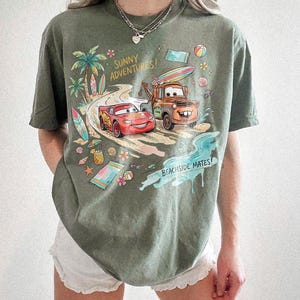 May include: An olive green short-sleeve t-shirt featuring a graphic print of Lightning McQueen and Mater from the movie Cars driving on a beach road. The text reads Sunny Adventures and Beachside Mates, surrounded by surfboards, shells, and flowers.