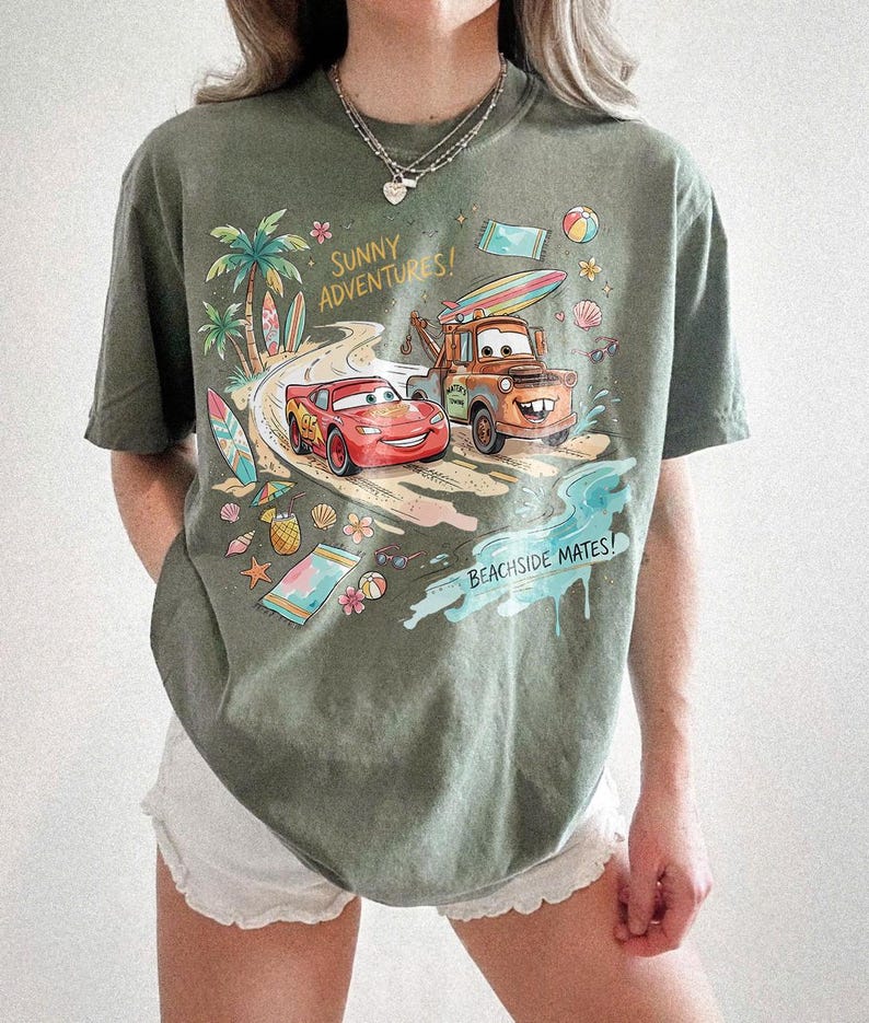 May include: An olive green short-sleeve t-shirt featuring a graphic print of Lightning McQueen and Mater from the movie Cars driving on a beach road. The text reads Sunny Adventures and Beachside Mates, surrounded by surfboards, shells, and flowers.