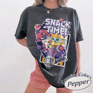 May include: A charcoal gray short-sleeve t-shirt featuring a graphic print with the text SNACK TIME! and Bounty Hunters. The design includes a green creature eating, surrounded by purple and pink clouds, and a helmet icon in a white frame.