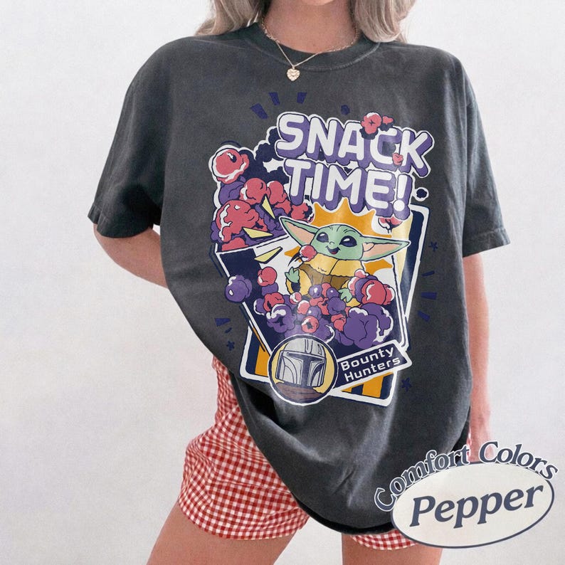 May include: A charcoal gray short-sleeve t-shirt featuring a graphic print with the text SNACK TIME! and Bounty Hunters. The design includes a green creature eating, surrounded by purple and pink clouds, and a helmet icon in a white frame.