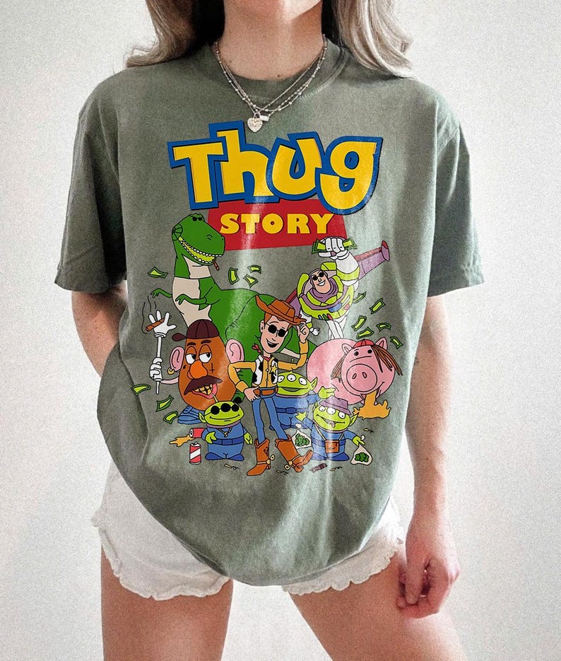 Pode incluir: Camiseta verde-oliva com um desenho de personagens de Toy Story e o texto "Thug Story". O design inclui um dinossauro, Woody, Buzz Lightyear e outros personagens de desenhos animados.