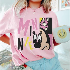 Op de afbeelding: Een lichtroze T-shirt met een Minnie Mouse-afbeelding en het woord "MINNIE" in roze, zwarte en gele blokletters. Een witte mok met een gele smiley wordt in de hand gehouden.